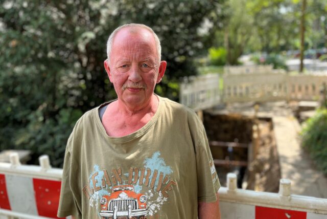 Ein Mann mit weit geschnittenem T-Shirt steht vor einer Baustelle. Der Gehweg hinter ihm ist aufgerissen und mit Baken abgesperrt.