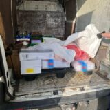 Die Ladefläche eines Transporters, in der sich Kisten befinden, die verpixelt sind. Eine Hand hält eine weiße Plastikplane nach oben, unter der sich ein Plastiksack befindet.