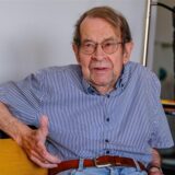 Ein älterer Mann mit Brille und etwas zersaustem Haar sitzt auf einer Ledercouch. Er trägt ein liniertes Hemd, das in die Jeanshose gesteckt ist.