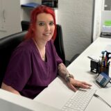 Sara Jane Bräunlich sitzt am Empfangstresen der Praxis Pelz und Weißbruch. Sie lächelt in die Kamera. Vor ihr steht der Arbeitscomputer, im Hintergrund ist ein Aktenschrank zu erkennen.