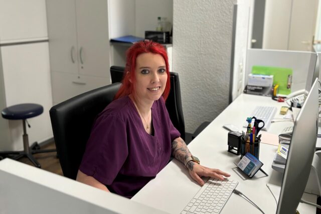 Sara Jane Bräunlich sitzt am Empfangstresen der Praxis Pelz und Weißbruch. Sie lächelt in die Kamera. Vor ihr steht der Arbeitscomputer, im Hintergrund ist ein Aktenschrank zu erkennen.