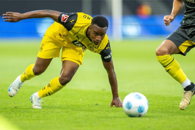 Youssoufa Moukoko stützt sich mit dem linken Arm auf dem Rasen ab und schaut auf den Ball.