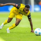 Youssoufa Moukoko stützt sich mit dem linken Arm auf dem Rasen ab und schaut auf den Ball.