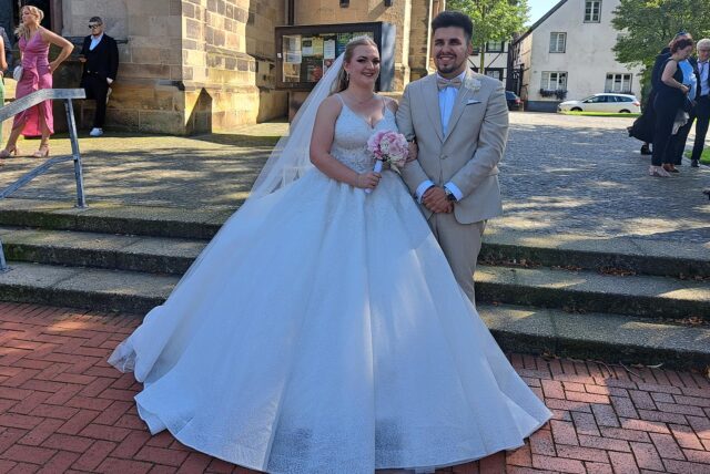 Michelle und Nino Mühlan stehen vor der St. Martinuskirche in Westerholt. Michelle trägt ein weißes Brautkleid, mit weitem Rock. Nino Mühan trägt einn Beigen Anzug mit gleichfarbiger Fliege.
