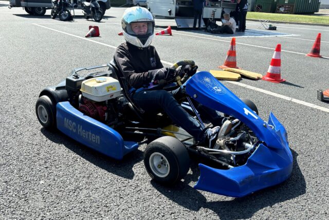 Maja Braun sitzt auf einem Blauen Kart mit der Aufschrift „MSC Herten“ auf der Seite. Maja Braun trägt einen weißen Helm mit dem Visier hochgeklappt und lächelt in die Kamera. Im Hintergrund stehen Hütchen ein Materialwagen und Motorroller. Davor stehen weitere Personen.