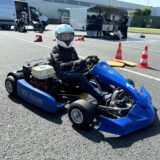 Maja Braun sitzt auf einem Blauen Kart mit der Aufschrift „MSC Herten“ auf der Seite. Maja Braun trägt einen weißen Helm mit dem Visier hochgeklappt und lächelt in die Kamera. Im Hintergrund stehen Hütchen ein Materialwagen und Motorroller. Davor stehen weitere Personen.