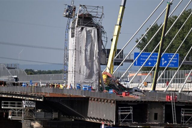 An einem alten Pfeiler-Stumpf, der für die Arbeiten mit grauer Plane eingehüllt ist, sieht man ein abgeknicktes verbogenes Baugerüst. Oben hängt ein schiefer Bauaufzug. Daneben steht ein gelber Kran.