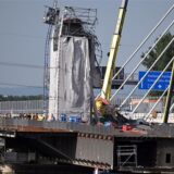 An einem alten Pfeiler-Stumpf, der für die Arbeiten mit grauer Plane eingehüllt ist, sieht man ein abgeknicktes verbogenes Baugerüst. Oben hängt ein schiefer Bauaufzug. Daneben steht ein gelber Kran.