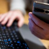 Eine Person hält ein Handy in der einen Hand und tippt mit der anderen Hand auf einer Computertastatur.