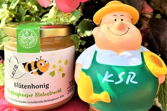 Auf einem gefüllten Honigglas steht Blütenhonig – Recklinghäuser Blütentracht. Daneben steht eine einem Gärtner nachempfundene Plastikfigur mit Sonnenhut, die eine Gießkanne und eine Sonnenblume hält. Auf der grünen Schürze steht KSR.
