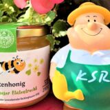 Auf einem gefüllten Honigglas steht Blütenhonig – Recklinghäuser Blütentracht. Daneben steht eine einem Gärtner nachempfundene Plastikfigur mit Sonnenhut, die eine Gießkanne und eine Sonnenblume hält. Auf der grünen Schürze steht KSR.