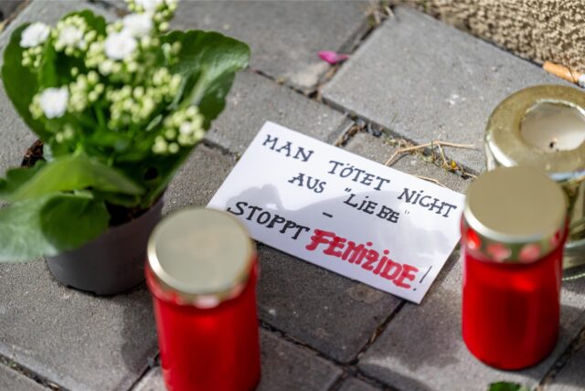 Blumen und Kerzen liegen vor einem Hauseingang neben einem Zettel mit der Aufschrift „Man tötet nicht aus Liebe – Stoppt Femizide“.