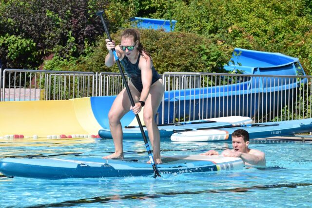 Eine Frau paddelt im Schwimmbecken auf einem Surfbrett und wird von einem Mann angeschoben.