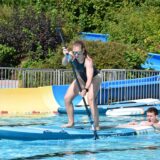 Eine Frau paddelt im Schwimmbecken auf einem Surfbrett und wird von einem Mann angeschoben.