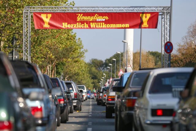 Die Kamera fotografiert zwischen zwei Reihen von Autos die Mittellinie einer Straße hinunter. Bei den Autos handelt es sich um Fahrzeuge aus dem Jahr 2004 bis 1994.Die Straße führt unter einem Banner her, auf dem in Gelb auf rotem Grund „Youngtimer Show Herzlichen Willkommen“ steht. Flankiert wird der Schriftzug von zwei gelben Y.