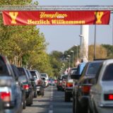 Die Kamera fotografiert zwischen zwei Reihen von Autos die Mittellinie einer Straße hinunter. Bei den Autos handelt es sich um Fahrzeuge aus dem Jahr 2004 bis 1994.Die Straße führt unter einem Banner her, auf dem in Gelb auf rotem Grund „Youngtimer Show Herzlichen Willkommen“ steht. Flankiert wird der Schriftzug von zwei gelben Y.