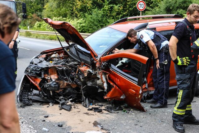 Hagen: Das Auto ist erheblich beschädigt. Polizeibeamte und Feuerwehrkräfte stehen neben dem zerstörten Auto.
