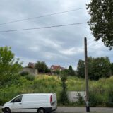 Ein Pkw steht auf einem Parkplatz am Straßenrand. Dahinter liegt eine verwucherte Grünfläche, die vom Bürgersteig mit einem Zaun getrennt ist. Hinter der Grünfläche stehen Wohnhäuser.