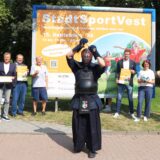 Ein Kendo-Kämpfer in schwarzer Schutzkleidung steht mit über dem Kopf erhobenen Stock vor einem großen Plakat, das die Verantaltung StadtSportVest 2024 in Recklinghausen ankündigt. Rechts und links der Werbetafel stehen acht an der Veranstaltung Beteiligte.