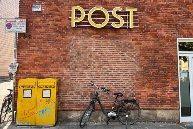 Ein Fahrrad steht vor einer Filiale der Postbank, die als Postfiliale fungiert, an der Backstein-Wand ist der Schriftzug «Post» zu lesen.