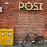 Ein Fahrrad steht vor einer Filiale der Postbank, die als Postfiliale fungiert, an der Backstein-Wand ist der Schriftzug «Post» zu lesen.