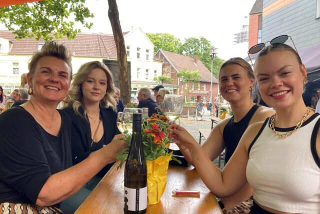 Vier Frauen sitzen an einem Biertisch und stoßen mit einem Glas Wein an.