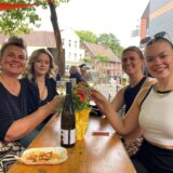 Vier Frauen sitzen an einem Biertisch und stoßen mit einem Glas Wein an.
