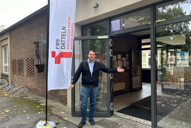 Simon Köcher steht neben einer Fahne mit FBS-Datteln-Logo und zeigt auf die offene Tür des Pfarrheim St. Josef.