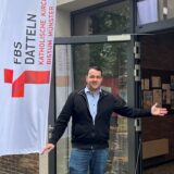 Simon Köcher steht neben einer Fahne mit FBS-Datteln-Logo und zeigt auf die offene Tür des Pfarrheim St. Josef.