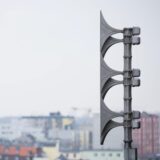 Eine Warn-Sirene steht im Vordergrund des Bildes. Im Hintergrund liegt verschwommen eine Stadtlandschaft mit mehreren Mehrfamilienhäusern und Wohnblöcken.