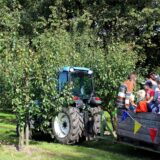 Ein kleiner Traktor fährt beim Erntedankfest in der Ried samt Anhänger durch eine Apfelplantage. Auf dem Anhänger ist eine große Kiste, in der Frauen und Kinder sitzen sowie stehen.