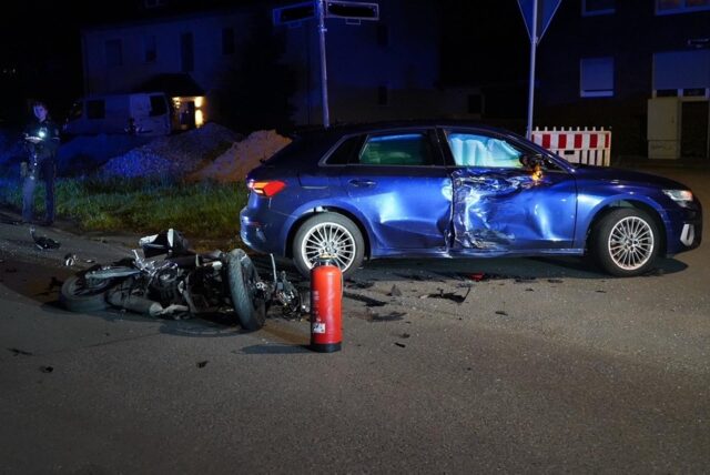 Motorradt liegt auf der Straße vor einem blauen Auto mit beschädigter Beifahrerseite.
