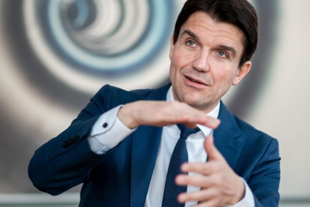Professor Uli Paetzel sitzt vor einer dekorierten Wand und gestikuliert.