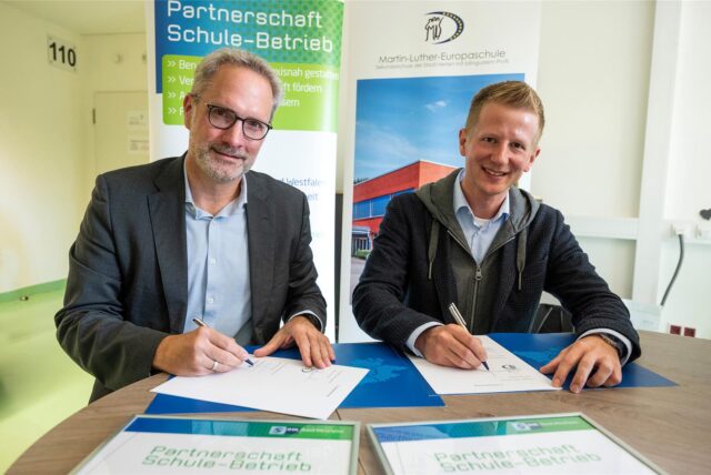 Thorsten Rattmann und Christoph Schenk sitzen an einem Tisch und unterzeichnen jeweils ein Papier, das vor ihnen liegt. Im Hintergrund weist ein Aufsteller auf das Projekt Partnerschaft Schule-Betrieb hin.