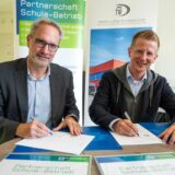 Thorsten Rattmann und Christoph Schenk sitzen an einem Tisch und unterzeichnen jeweils ein Papier, das vor ihnen liegt. Im Hintergrund weist ein Aufsteller auf das Projekt Partnerschaft Schule-Betrieb hin.