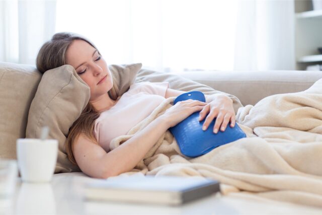 Eine Frau liegt auf einem Sofa, auf ihrem Bauch liegt eine Wärmflasche.