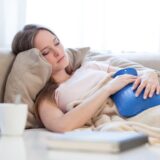 Eine Frau liegt auf einem Sofa, auf ihrem Bauch liegt eine Wärmflasche.