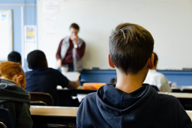 Ein Schüler (von hinten) lauscht den Ausführungen des Lehrers im Hintergrund.