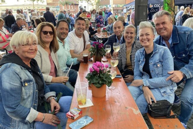 Eine Gruppe von Menschen sitzt an einem Tisch im Freien und genießt Getränke. Im Hintergrund sind weitere Menschen und Gebäude zu sehen.