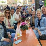 Eine Gruppe von Menschen sitzt an einem Tisch im Freien und genießt Getränke. Im Hintergrund sind weitere Menschen und Gebäude zu sehen.