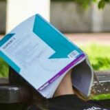 Links im Bild ein Jugendlicher, der mit einem Schulheft auf dem Gesicht in einem Park schläft. Rechts im Bild ein Wecker, der 8.05 Uhr anzeigt.