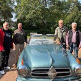 Vier Männer stehen auf einem Ascheparkplatz an einem Oldtimer, zwei auf jeder Seite.