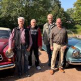 Vier Männer stehen auf einem Ascheparkplatz zwischen zwei Oldtimern.