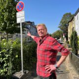 Ein Mann mit rot kariertem Hemd steht auf einem Bürgersteig und zeigt auf ein Halteverbotsschild.