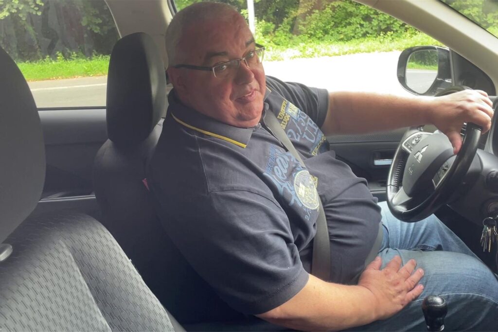 Ein Mann sitzt hinter dem Steuer seines Autos und blickt in die Kamera. Ein Mann sitzt hinter dem Steuer seines Autos und blickt in die Kamera.