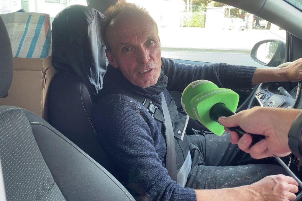 Ein Autofahrer sitzt am Steuer und wird von einem Reporter befragt. Man sieht ein Mikrofon mit der Aufschrift Ein Autofahrer sitzt am Steuer und wird von einem Reporter befragt. Man sieht ein Mikrofon mit der Aufschrift