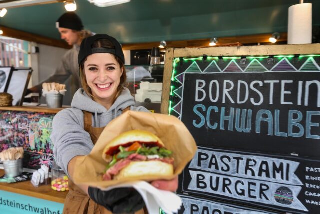 Eine Gastronomin zeigt einen Pastrami-Burger in die Kamera.