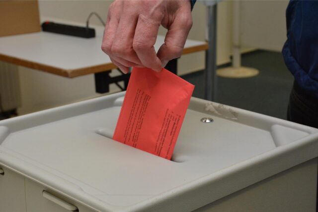Eine Hand in Großaufnahme wirft einen roten Wahlbrief-Umschlag in eine Wahlurne.