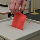 Eine Hand in Großaufnahme wirft einen roten Wahlbrief-Umschlag in eine Wahlurne.