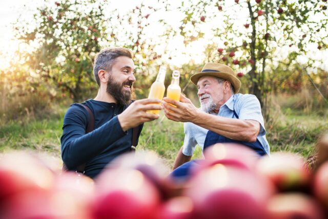 Zwei Männer sitzen vor Apfelbäumen und stoßen mit Apfelsaft an.